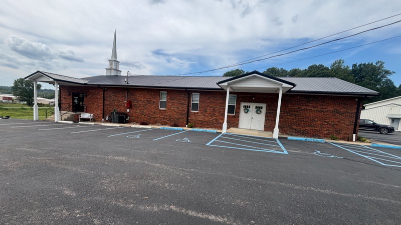 New Beginning Church of the Nazarene seen from its car park: a long red-brick building with a white steeple, a covered porch over white double doors toward the right, and marked accessible parking bays directly in front of them.